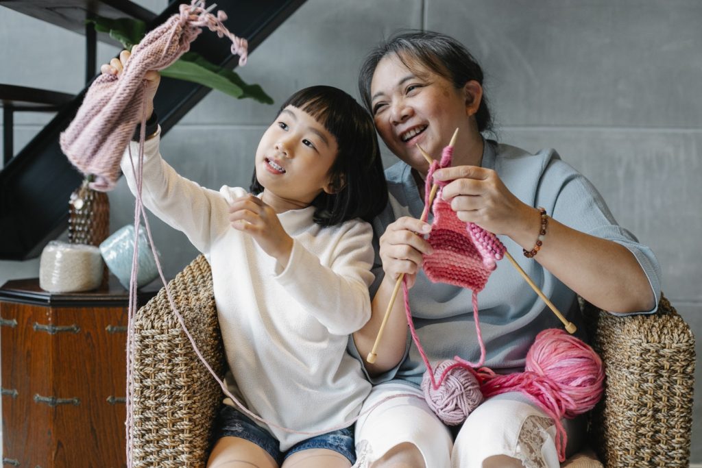 Happy Asian girl showing knitting to laughing grandmother both sitting in cozy armchair