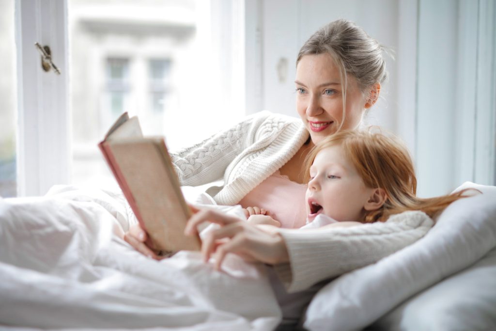 Cheerful young woman hugging cute little girl and reading book together while lying in soft bed in light bedroom at home in daytime