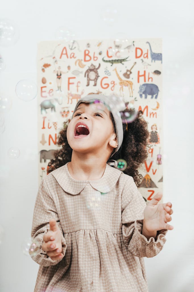 A happy child laughing while playing with bubbles against a colorful alphabet backdrop indoors.