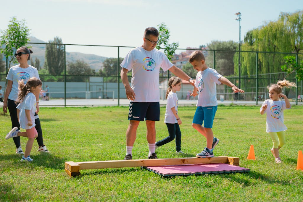 Kids guided by instructors at a park balance on a beam for fun and exercise.